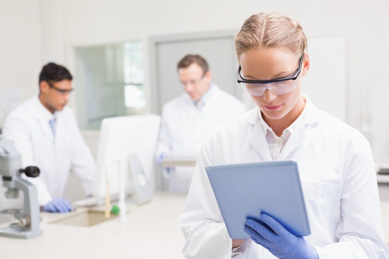 Female scientist, wearing goggles, using tablet while colleagues are working behind her