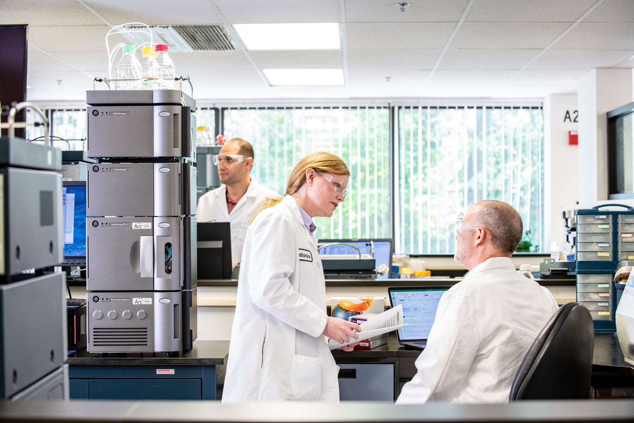 Trois scientifiques portant des lunettes de protection dans un laboratoire lumineux. Deux d’entre eux communiquent à côté d’un système Arc Premier.