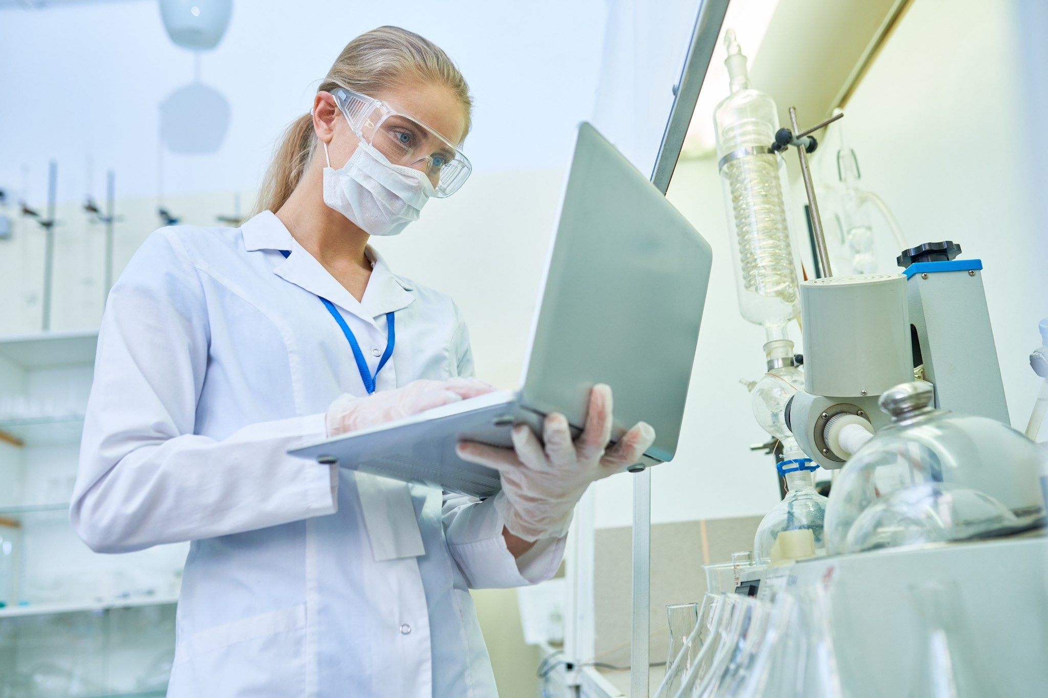 Une jeune scientifique sérieuse et sûre d’elle, portant un masque de protection et des lunettes de sécurité, examine le fichier contenant les résultats d’une expérience sur un ordinateur portable, tout en travaillant avec du matériel médical